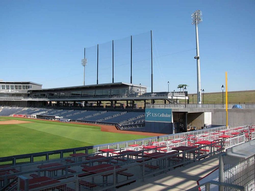 An large baseball stadium with bleacher seating and sky boxes above, green field, and tall floodlights under a blue sky. There is a tall netting system behind the sky boxes protecting the parking lot from stray balls. 