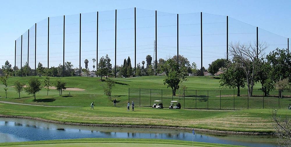 Golfers enjoy a lush green golf course by a serene pond, with an advanced golf course netting system standing tall in the background. Two golf carts rest nearby under the clear blue sky.