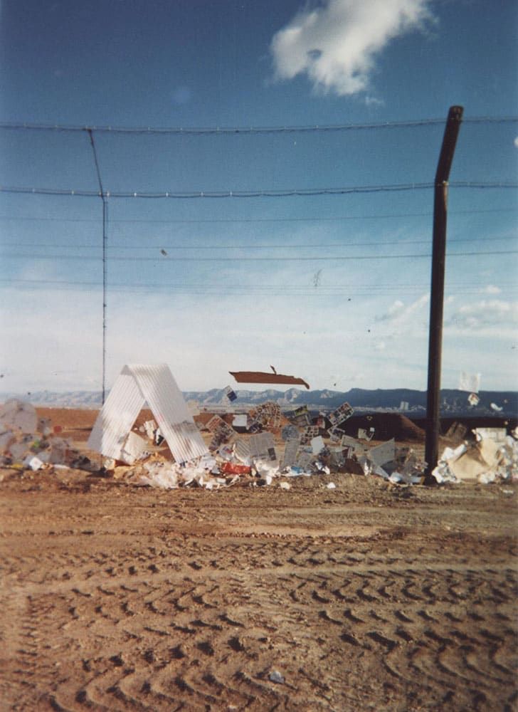 A pile of trash and debris piles up at the base of a stationary litter fence. Tire tracks are visible in the foreground. There are clear skies in the background. 