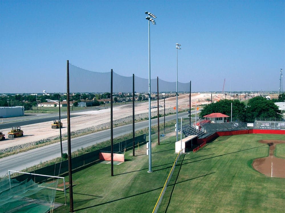 A baseball field with bleachers, with a tall netting fence between the field and a road to the left. There is another road under construction on the side, with several construction vehicles visible.