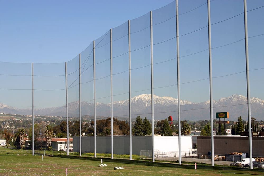 A driving range with tall netting in the foreground, and distant mountains topped with snow under a clear blue sky in the background. Several buildings and trees are also visible.
