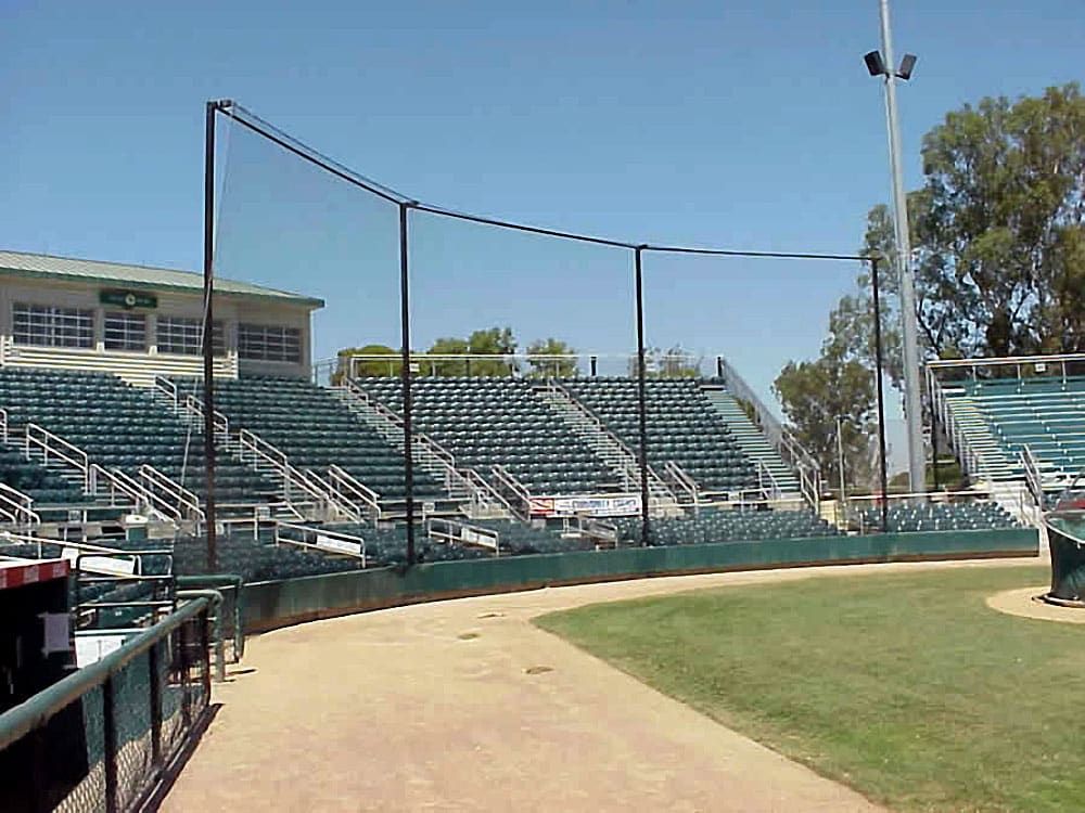 Baseball stadium with empty green and white bleachers, protective netting fence, and a well-maintained grass field under a clear blue sky.