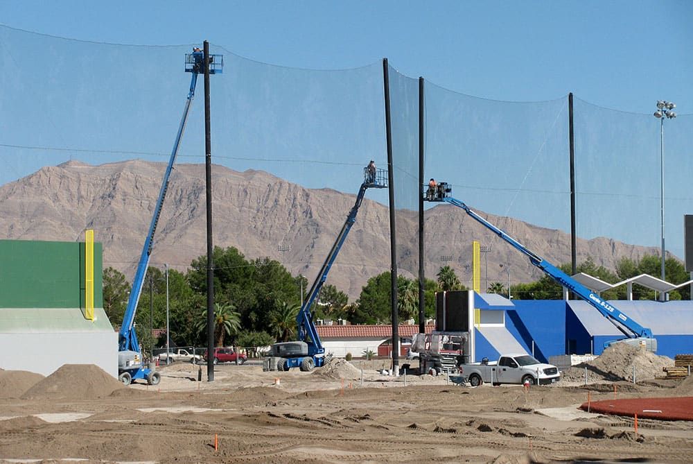 Three blue construction lifts are used by workers to erect tall black netting poles on a sports complex construction site. Vehicles and building materials are scattered around, with a dry mountain in the background.