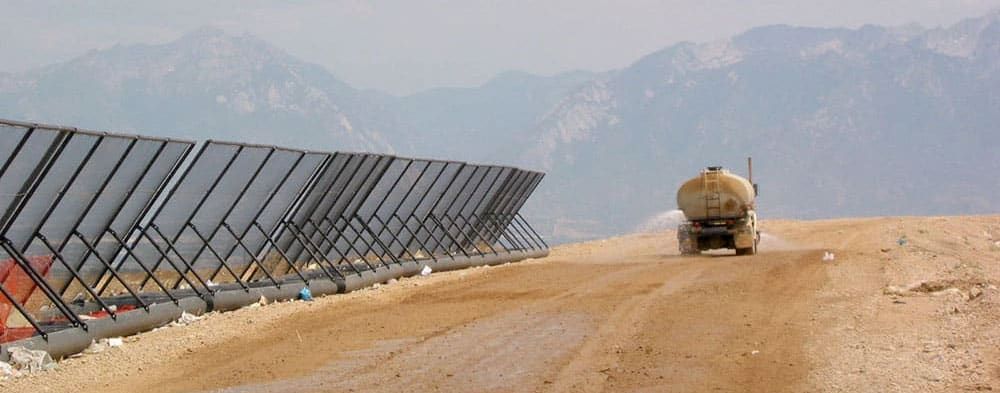 A truck sprays water on a dirt road beside rows of portable litter fences with mountains in the background.