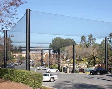A parking lot with several cars is surrounded by tall poles supporting a large, netting structure overhead, protecting the cars from errant golf balls. Trees and buildings are visible in the background.