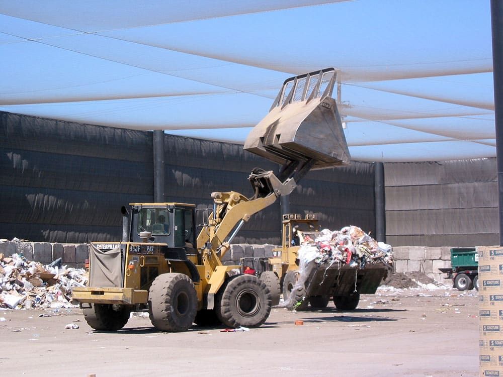 Front loader tractors move scoops of mixed waste, with piles of sorted materials in the background. The recycling center is enclosed with full overhead litter containment netting and privacy netting walls.