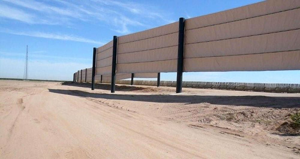 A tall, long border wall constructed with poles and tan fabric material provides visual blockage of what’s inside.