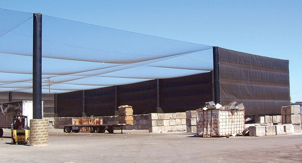 A transfer station enclosed with full overhead litter containment netting and privacy netting walls. There are stacked blocks, a forklift and a trailer with boxed items on a concrete floor.