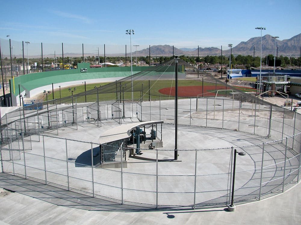 Outdoor baseball and softball batting cages with nets around and above. and lights, adjacent to a green baseball field with tall ights and red infield, surrounded by a chain-link fence. Nearby are various sports complex facilities and mountainous landscape.