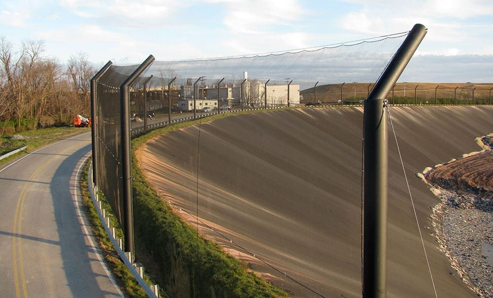 A curved road runs alongside a secured industrial facility with high stationary litter fencing. There is a backdrop of barren trees and construction equipment.