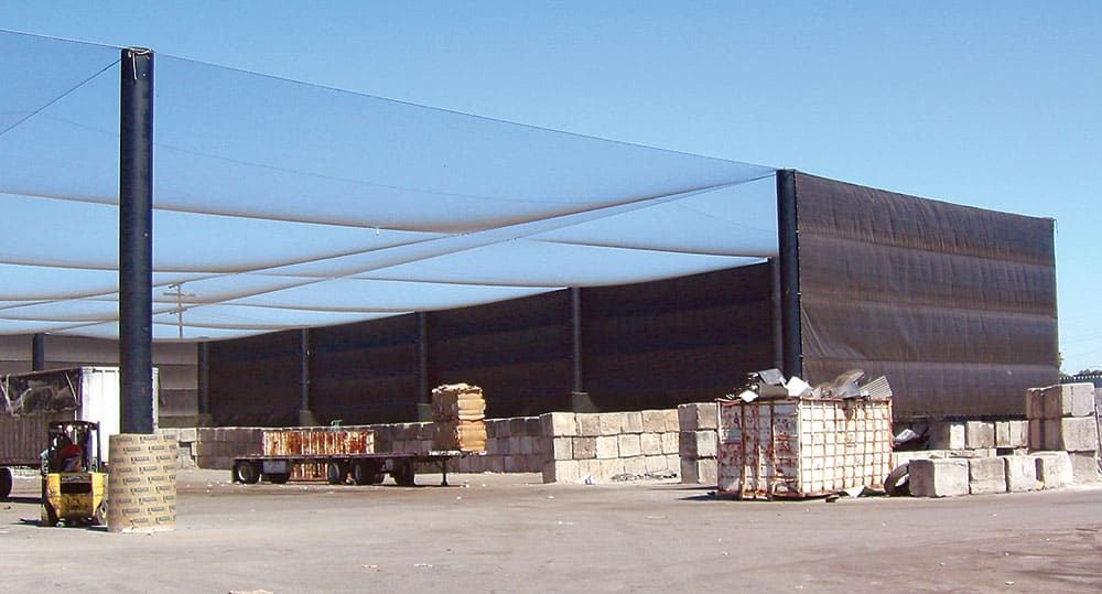 A large transfer station covered and surrounded by netting and tall poles. A forklift and stacks of boxes, pallets, and other refuse are visible on the ground.