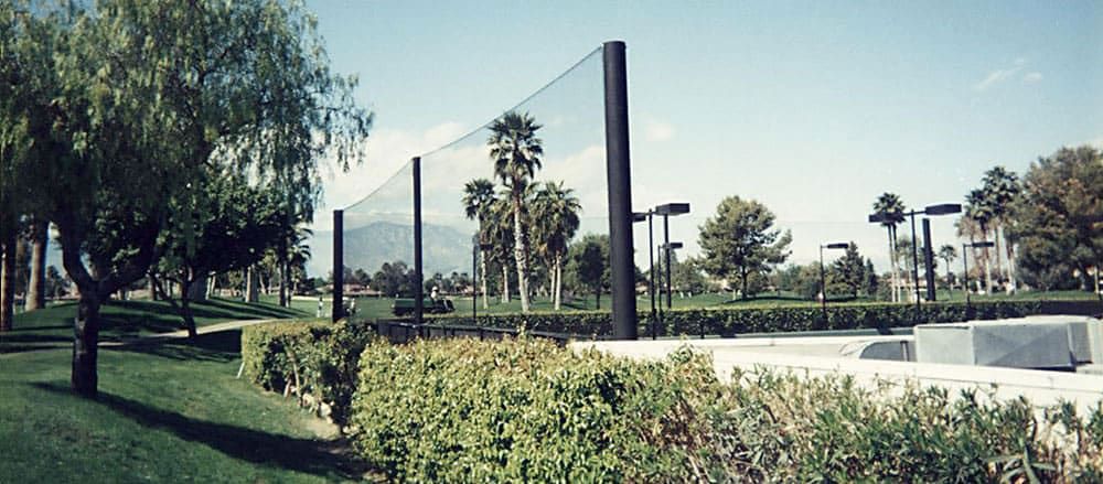 Tennis court with netting system in a lush green park landscape with palm trees and bushes. Mountains are visible in the background under a clear blue sky.