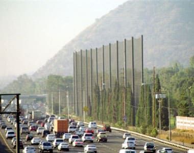 Busy freeway with multiple lanes of traffic passing by a large golf net installation set against a hilly background.