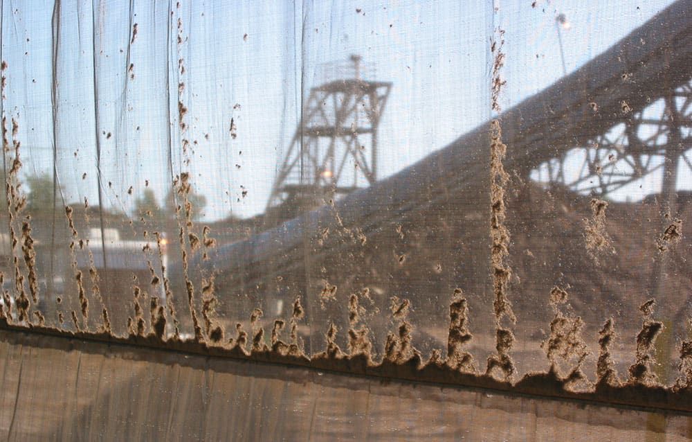 Close-up of a windscreen collecting a lot of dust, with a blurred view of industrial structures and a blue sky in the background.