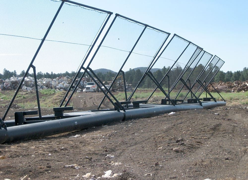 Five Coastal Netting portable litter fences aligned in a dirt field. 2 large trucks are parked in the background next to a largepile of trash inside a fence. Mountains and trees are visible beyond.