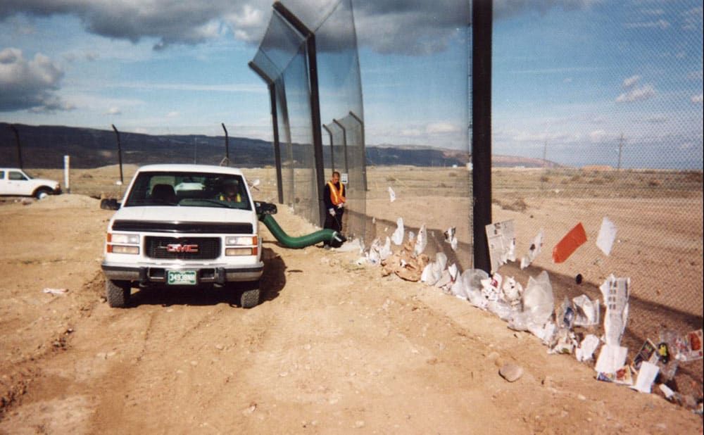 A white GMC pickup with a green hose sucks up lots of trash collected at the base of a stationary litter fence in a barren landscape. A worker in an orange vest stands nearby.