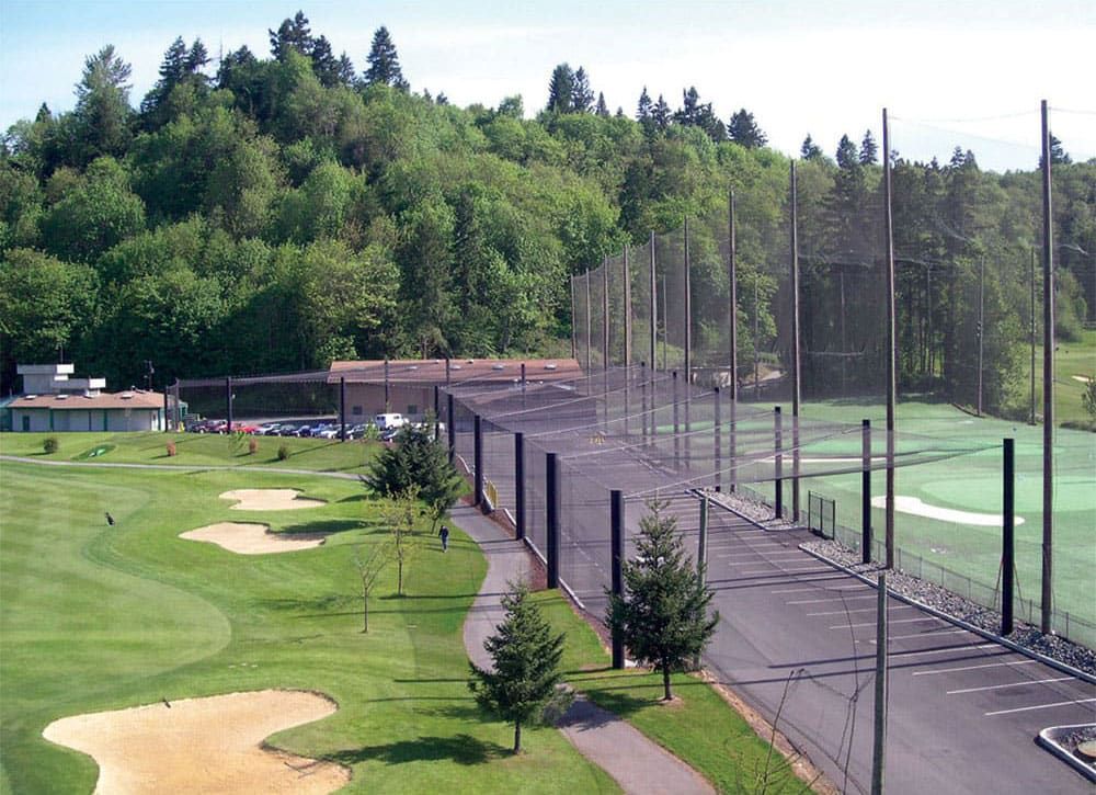 A driving range with protective netting on the right and a landscaped golf course with sand bunkers and putting greens on the left and parking lot on the right. A wooded hillside and buildings are in the background.