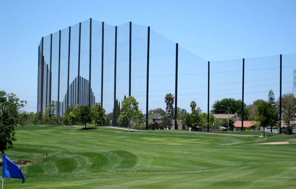 Golf course with tall black netting poles in the background, blue flag on a foreground green. Trees and houses are visible beyond the netting. Clear blue sky.