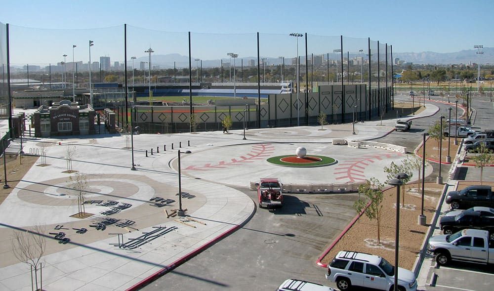 An elevated view of an urban sports complex featuring a baseball field surrounding by a tall netting system, a circular plaza with a central sculpture, and parking lots.